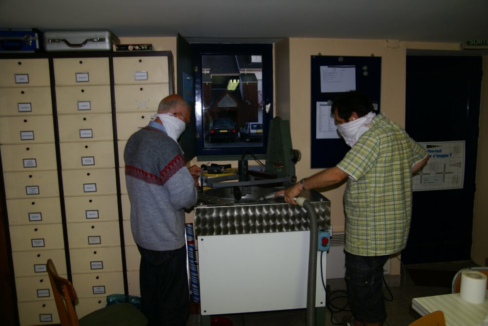 En soirée dans un local, deux personnes avec un foulard sur le nez s'affairent autour de la machine à tailler les miroirs qui produit visiblement beaucoup de poussières fines... et du bruit.