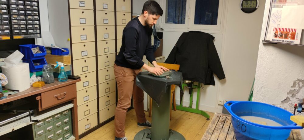 Bryan derrière le pied colonne en train frotter le miroir sur un disque outil  en verre. 