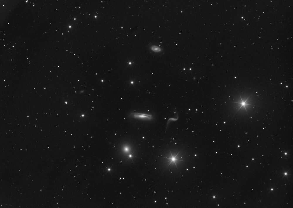 Image monochrome d'un amas de galaxies dans la constellation du Lion. Proche du centre une fine galaxie en forme d'intégrale mathématique. Sur sa gauche, une galaxie spirale traversée d'un disque de poussière. En haut de l'image au centre, une petite galaxie spirale barrée en forme de volant. En bas de l'image à gauche, près du centre se trouve une galaxie elliptique de forme circulaire, à proximité d'une étoile. D'autres petites taches floues également réparties sur l'image pour autant de faibles galaxies. Dans le quart bas et droite de l'image se trouve deux étoiles brillantes à six branches. Les branches sont le résultat de l’artefact produit par le support du miroir secondaire (appelé araignée) du télescope qui a permis la prise de vue. Les trois branches de l'araignée produisent par un effet miroir les six branches des étoiles. D'autres étoiles plus faibles ne produisant pas d'artefacts complètent l'image.