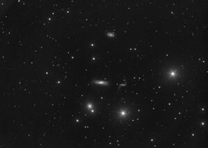 Image monochrome d'un amas de galaxies dans la constellation du Lion. Proche du centre une fine galaxie en forme d'intégrale mathématique. Sur sa gauche, une galaxie spirale traversée d'un disque de poussière. En haut de l'image au centre, une petite galaxie spirale barrée en forme de volant. En bas de l'image à gauche, près du centre se trouve une galaxie elliptique de forme circulaire, à proximité d'une étoile. D'autres petites taches floues également réparties sur l'image pour autant de faibles galaxies. Dans le quart bas et droite de l'image se trouve deux étoiles brillantes à six branches. Les branches sont le résultat de l’artefact produit par le support du miroir secondaire (appelé araignée) du télescope qui a permis la prise de vue. Les trois branches de l'araignée produisent par un effet miroir les six branches des étoiles. D'autres étoiles plus faibles ne produisant pas d'artefacts complètent l'image.