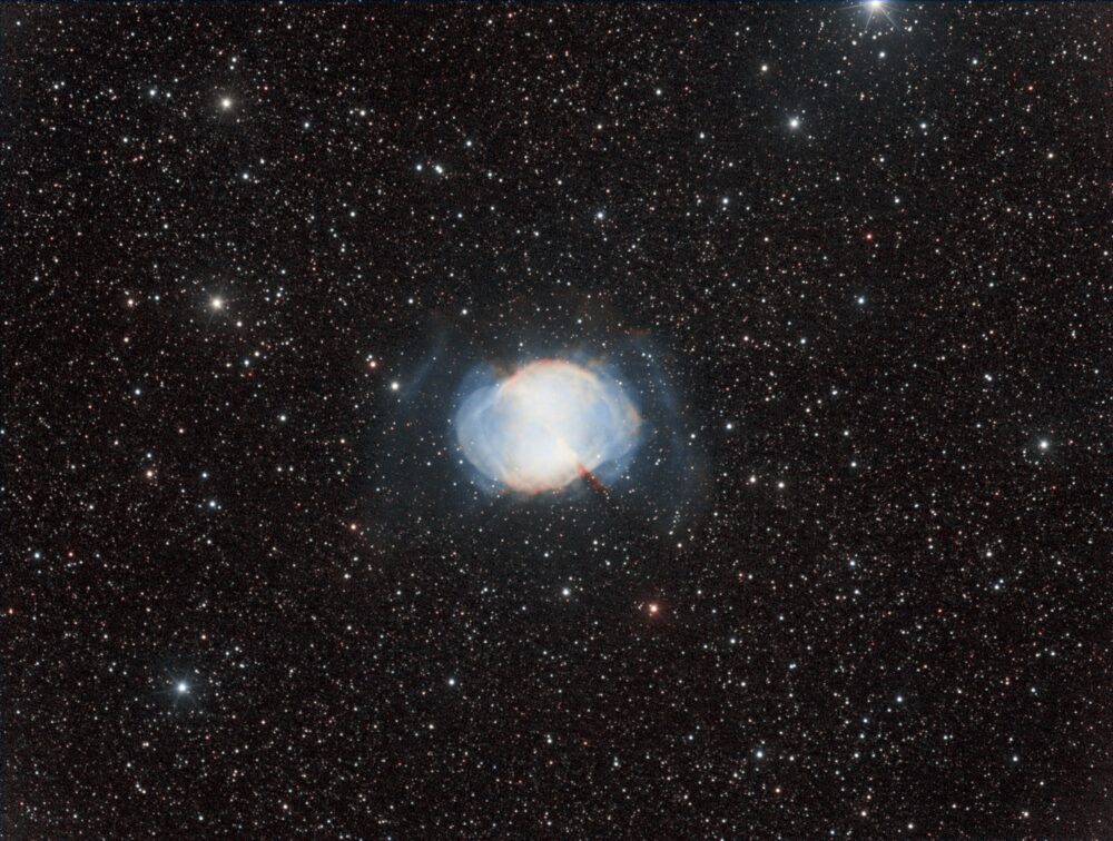 Nébuleuse planétaire de l'Haltère au centre de l'image. De forme ovale, un peu comme un œuf couché, de teinte blanche tirant sur le bleuen s'éloignant du centre saturé. Quelques volutes bleues beaucoup moins denses en forme de parenthèses apparaissent de part et d'autre de la nébuleuse. Un filament de gaz de couleur rouge apparaît en périphérie de la nébuleuse sur un axe 10h/4h. Le fond de ciel est modérément dense en étoiles